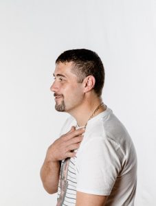 Side profile of a man with short dark hair and goatee, wearing a white t‑shirt and chain, with one hand on his chest.