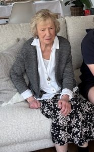 Elderly woman with light hair sitting on a light sofa, wearing a white blouse, gray cardigan, and a floral skirt in a living room.