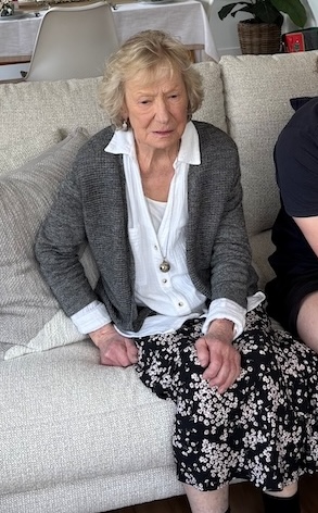Elderly woman with light hair sitting on a light sofa, wearing a white blouse, gray cardigan, and a floral skirt in a living room.