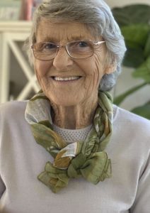 Smiling elderly woman with short gray hair and glasses wearing a green patterned scarf indoors.