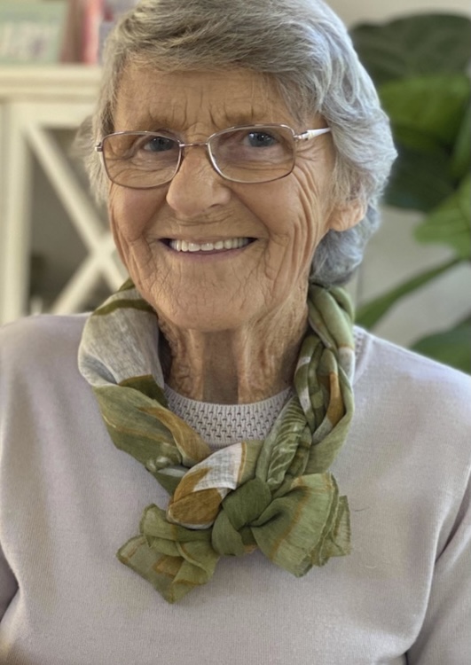 Beryl Woodcock Smiling elderly woman with short gray hair and glasses wearing a green patterned scarf indoors.