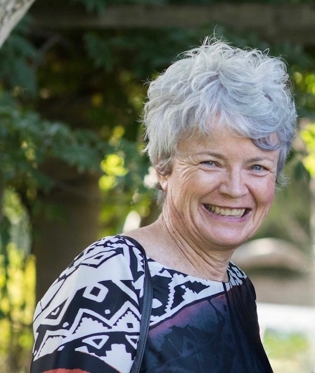 Catherine Dobson Smiling older woman with short gray hair wearing a patterned top outdoors with greenery in the background.