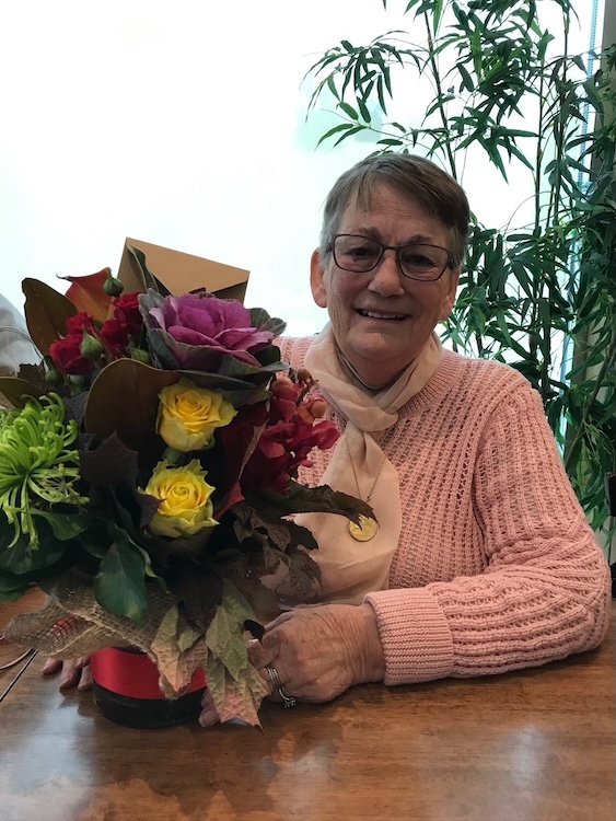 Christina SECKOLD Smiling elderly woman in a pink sweater sits at a wooden table, holding a colorful bouquet of yellow and red flowers with green leaves, in front of a potted plant.