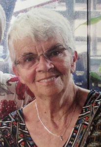 Smiling older woman with short white hair and glasses, wearing a patterned blouse and a silver necklace, indoors near a glass window.