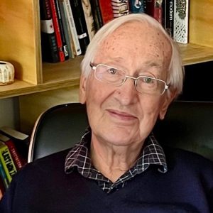 Older man with white hair and glasses sits in a chair, wearing a navy sweater over a checkered shirt, in front of a bookshelf.