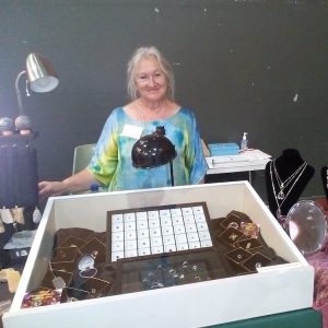 Smiling jewelry vendor standing behind a glass display case filled with rings and necklaces, at a market stall.
