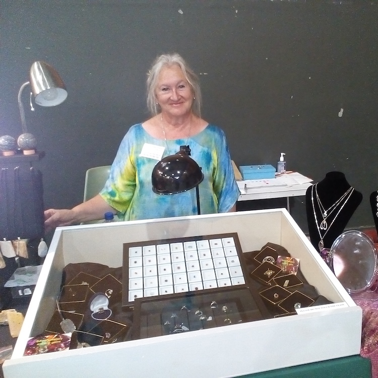 Helen Butrumlis Smiling jewelry vendor standing behind a glass display case filled with rings and necklaces, at a market stall.