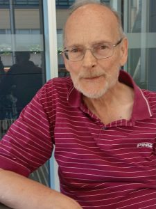 Older man with glasses and light beard sitting outdoors beside a glass building, wearing a maroon striped polo.