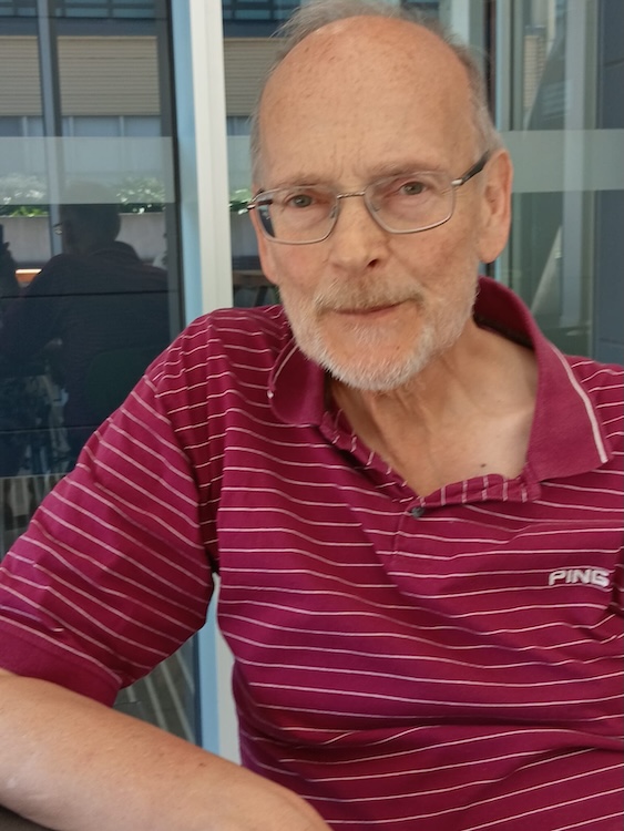 Older man with glasses and light beard sitting outdoors beside a glass building, wearing a maroon striped polo.