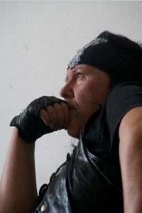 Man in a bandana and black leather vest rests his chin on a gloved hand, looking thoughtful in profile.