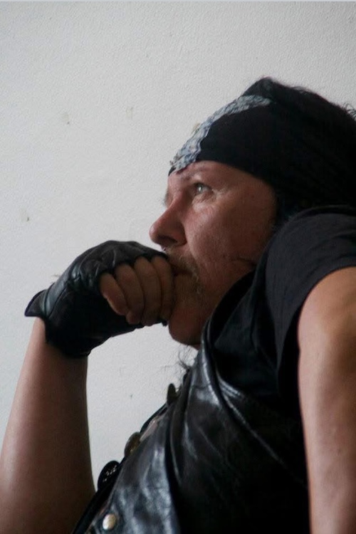 Man in a bandana and black leather vest rests his chin on a gloved hand, looking thoughtful in profile.