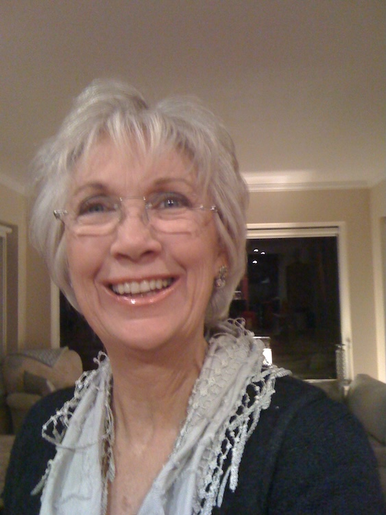 Smiling older woman with short gray hair and glasses, wearing a light scarf, indoors in a living room.