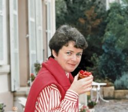 Woman with short dark hair smiling, wearing a red vest over a striped shirt, holding a cupcake with red icing on a cobblestone street outside a café.