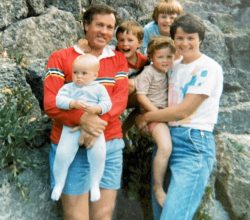 Family of six posing on a rocky outdoor hillside; two adults hold children while three kids stand and smile behind them.
