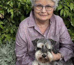 Smiling elderly woman with blue glasses holding a small Schnauzer outdoors, green hedge behind them.
